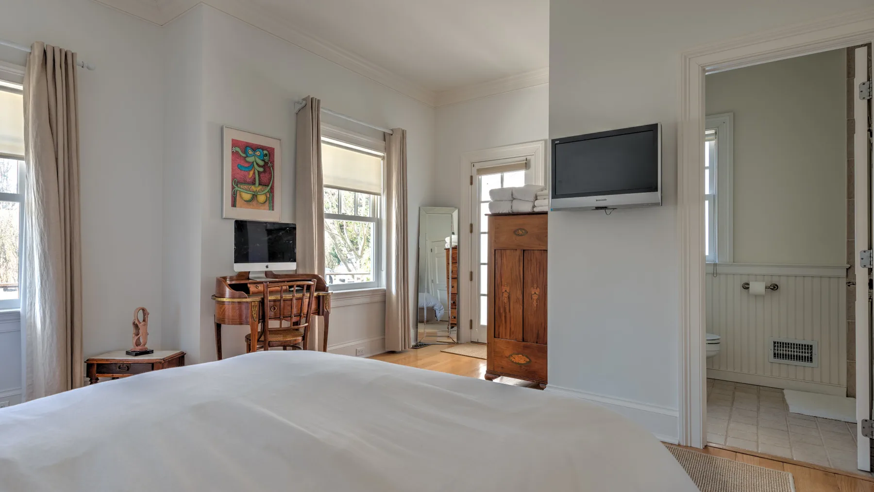A bedroom with a neatly made bed, tall windows, and a wall-mounted television.