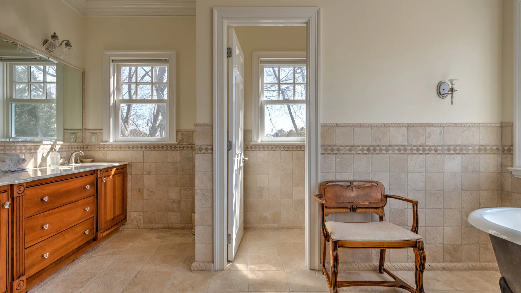 A tiled bathroom with a wooden vanity, a chair, and windows.