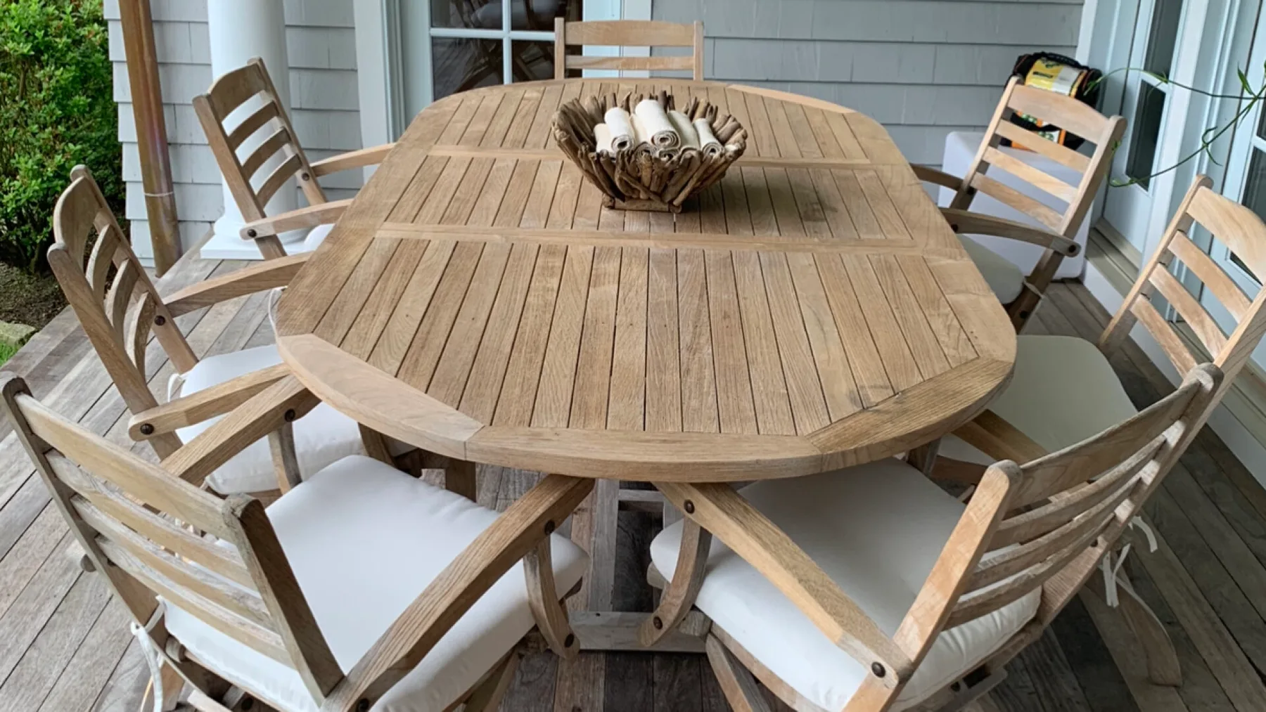 A wooden outdoor dining table is surrounded by cushioned seating on a covered patio.
