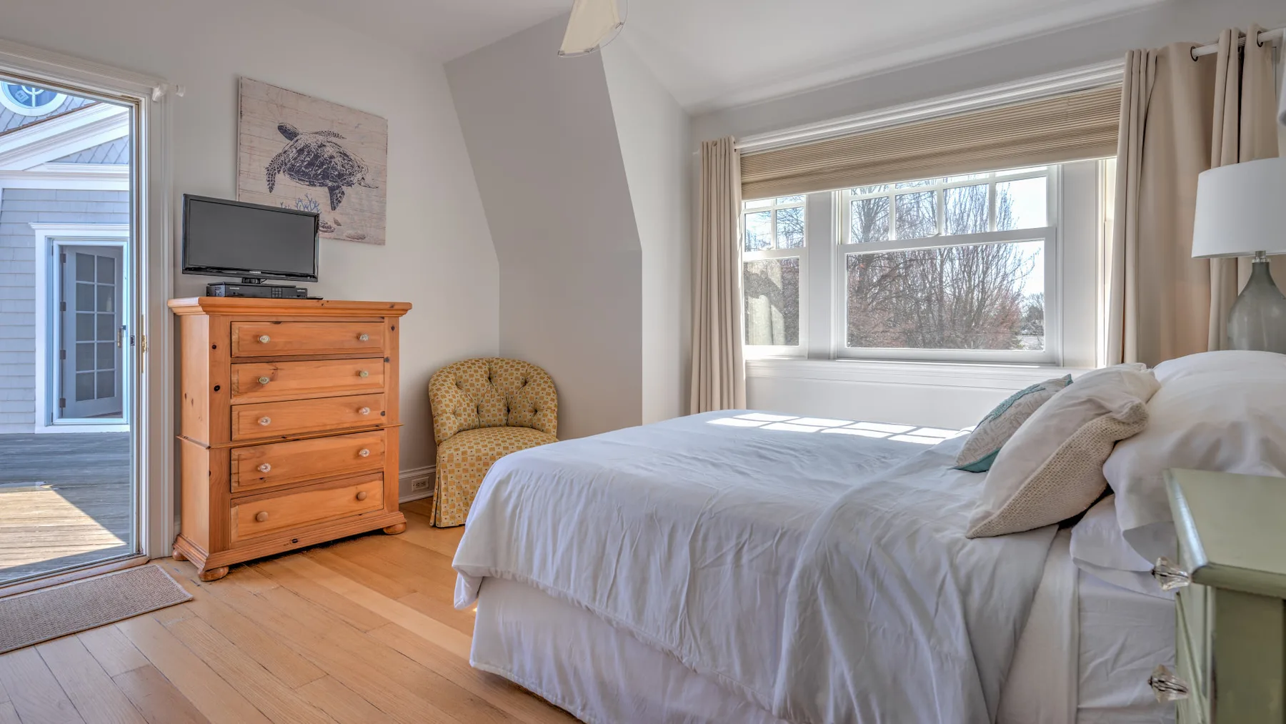 A bright bedroom with a bed, wooden dresser, and windows overlooking trees.