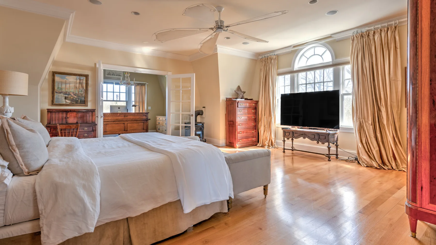 A bright bedroom with a large bed, a television, and tall curtained windows.