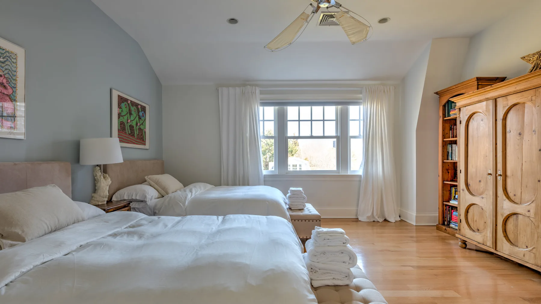A bright bedroom with two beds, a window, and a wooden wardrobe.