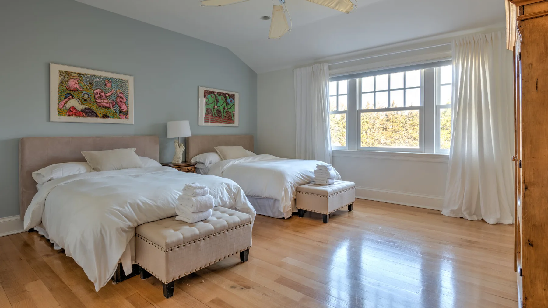 A bright bedroom with two beds, a large window, and light wood flooring.