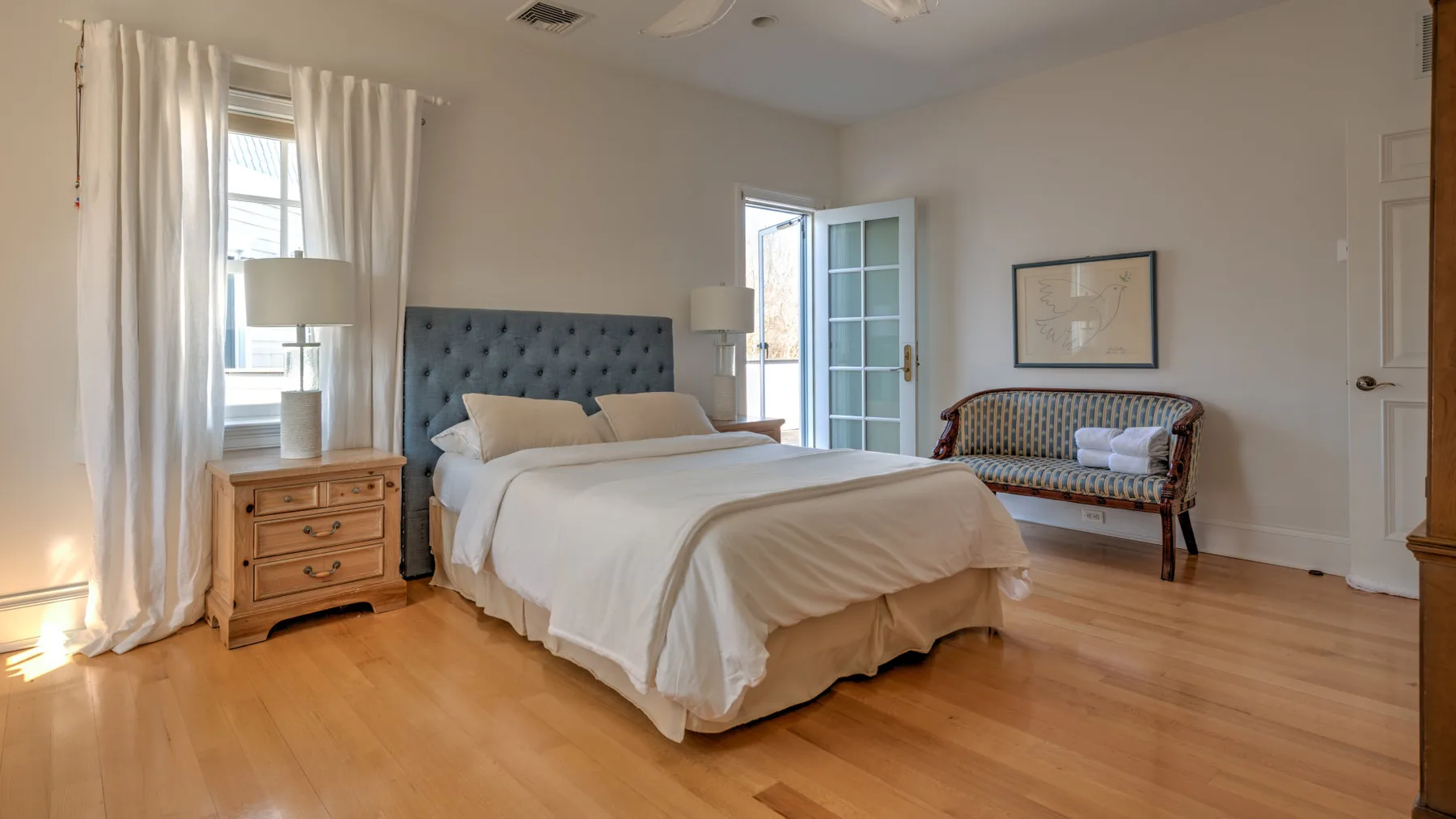 A bright bedroom with a large bed, bedside table, and tall windows.