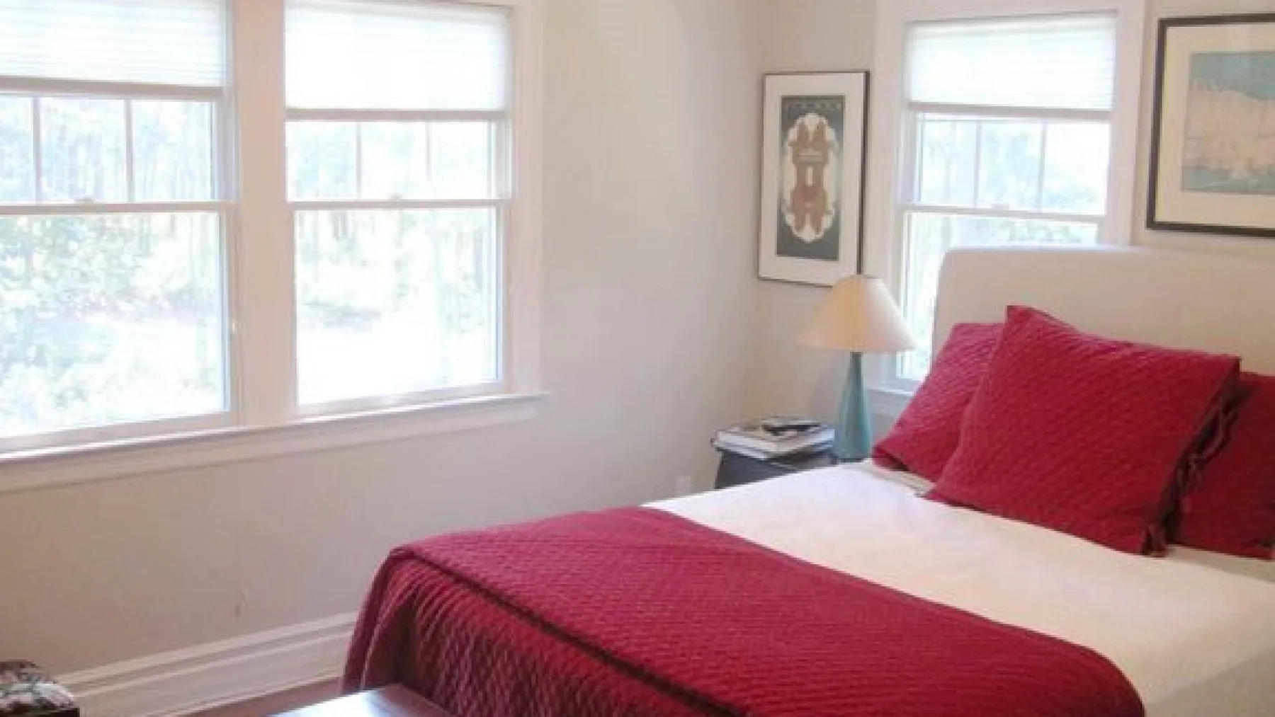 A bedroom with a bed dressed in red bedding beside several windows.