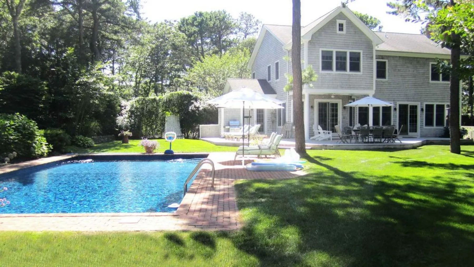 A backyard pool beside a large gray house, with patio seating and trees.