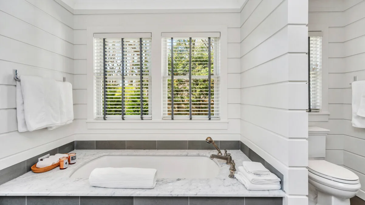 Un baño luminoso con una bañera bajo ventanas con persianas.