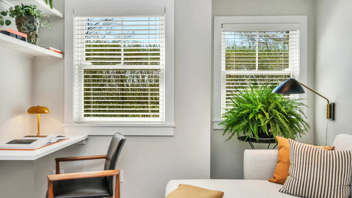 Una habitación luminosa con escritorio, persianas en las ventanas y asientos junto a una planta frondosa.