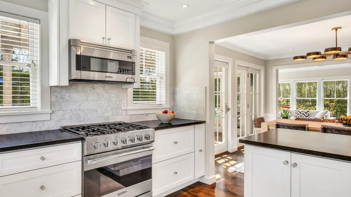 Una cocina luminosa con gabinetes blancos y una estufa se abre a un comedor soleado.