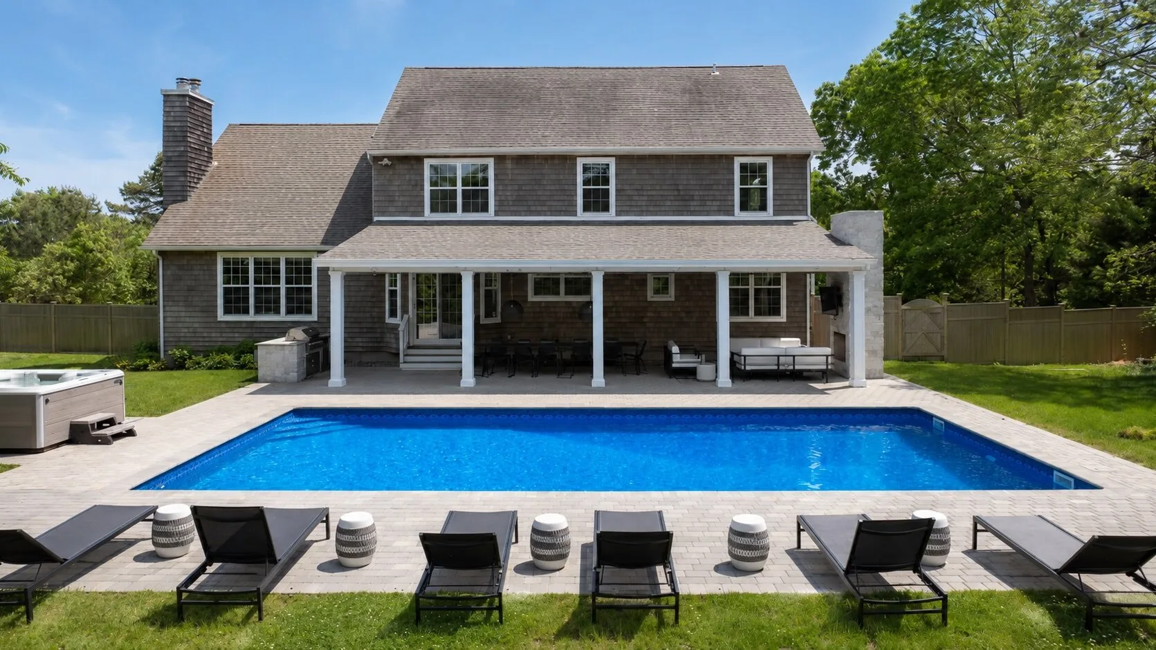 A backyard swimming pool sits beside a large house, with lounge chairs along the deck.