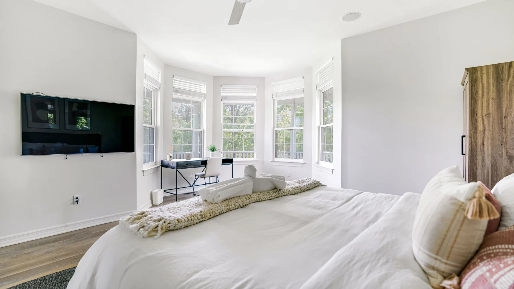 A bright bedroom with a large bed, a wall-mounted television, and several windows.