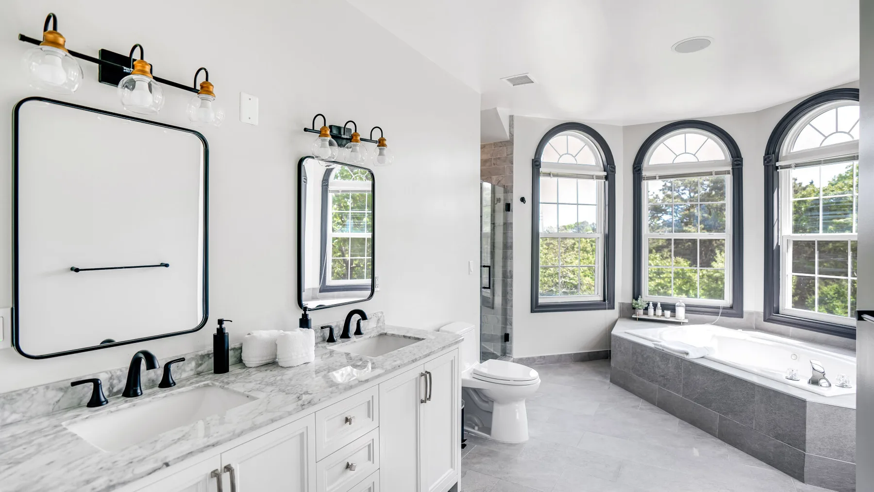 A bright bathroom has a long vanity, mirrors, and a bathtub beneath arched windows.
