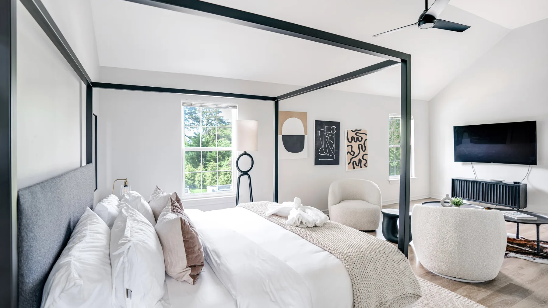 A bright bedroom with a large bed, pale seating, and tall windows.