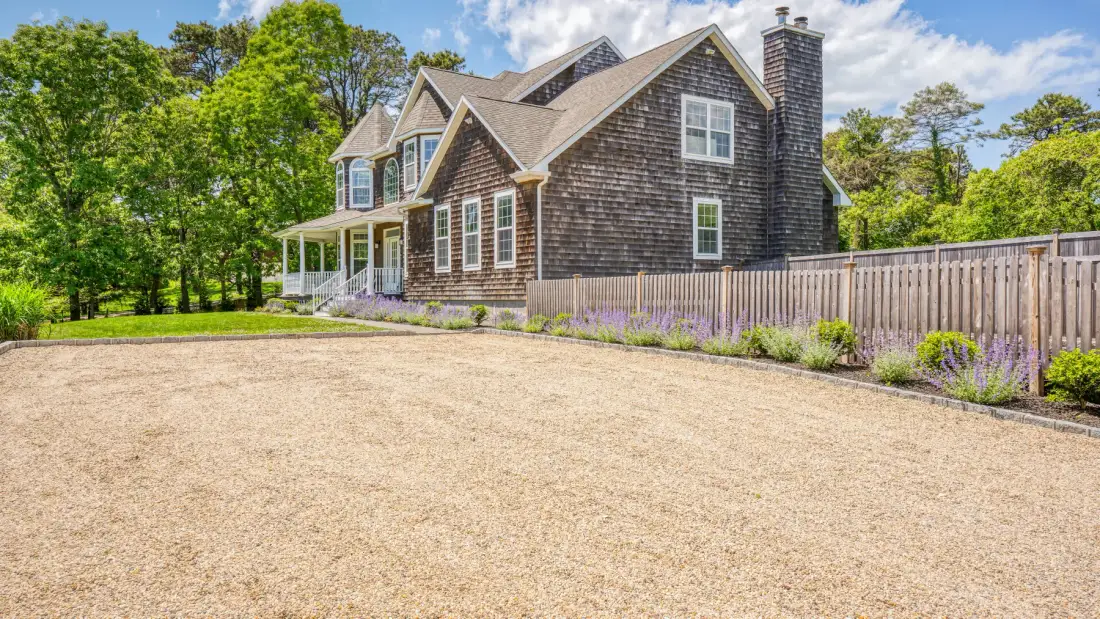 A large shingled house stands beside a gravel driveway and wooden fence.