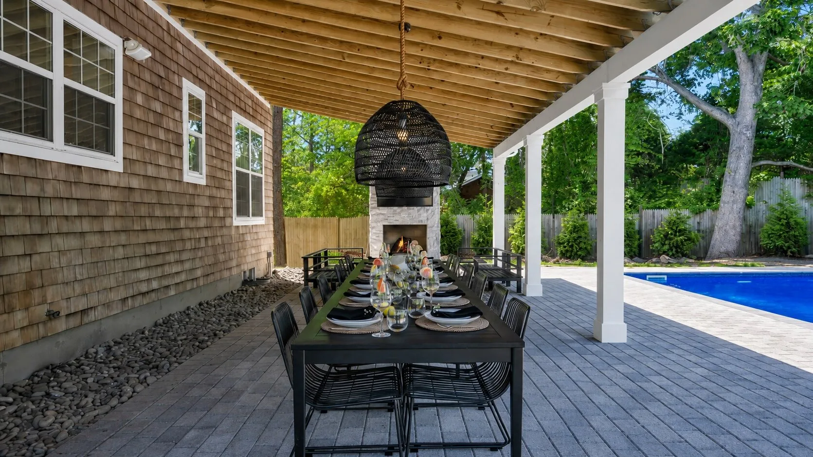 A covered outdoor dining area has a long table set beside a swimming pool.