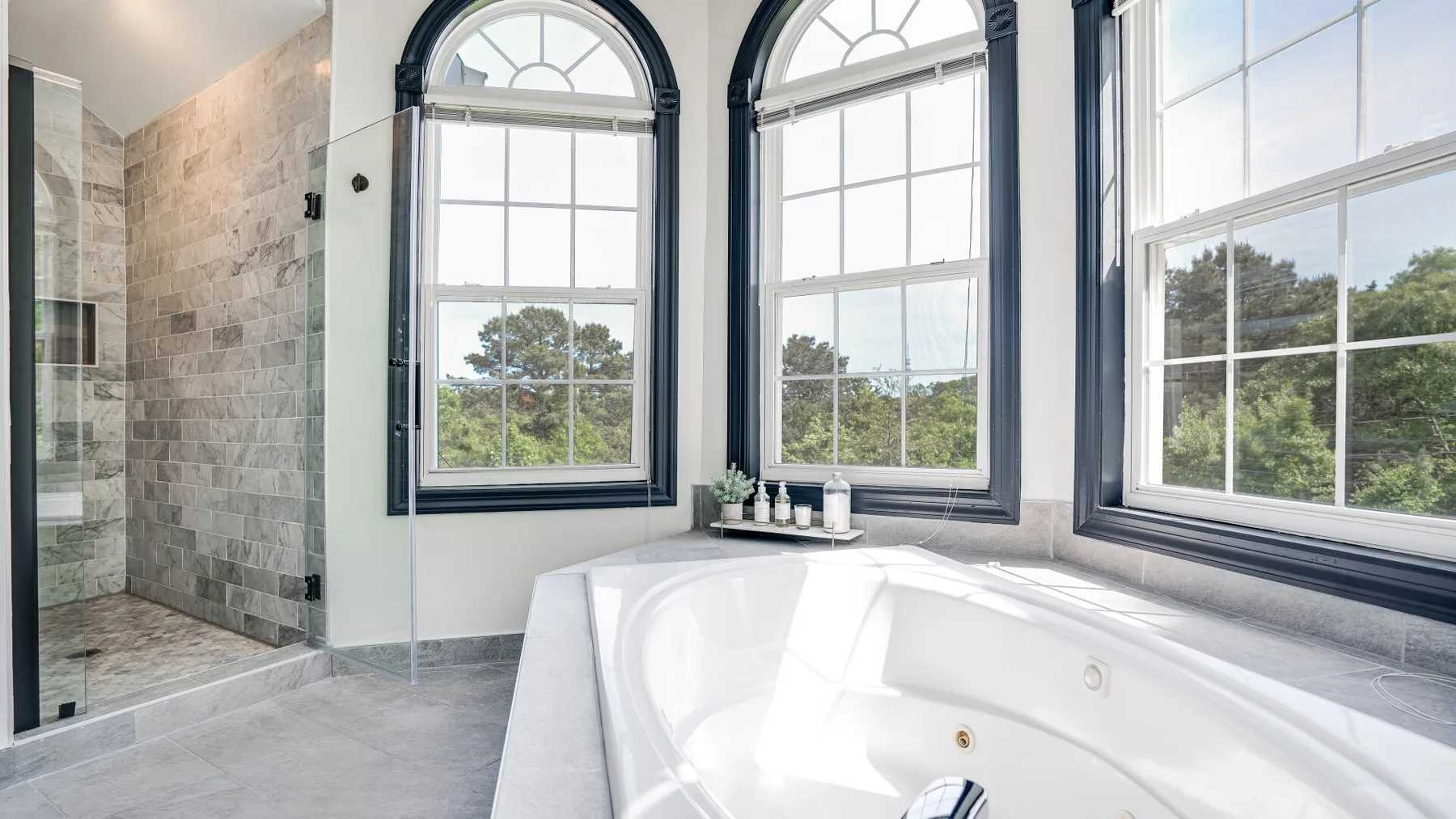 A bright bathroom features a large bathtub beside tall windows and a glass shower.