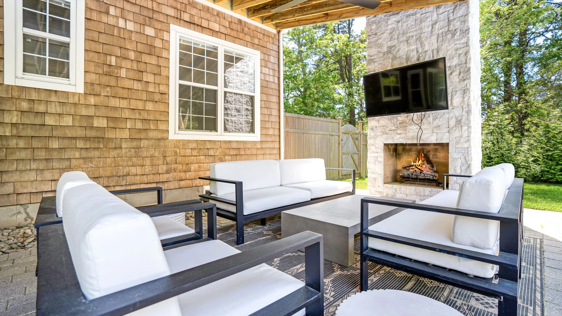 An outdoor seating area with a stone fireplace and television beneath a covered patio.