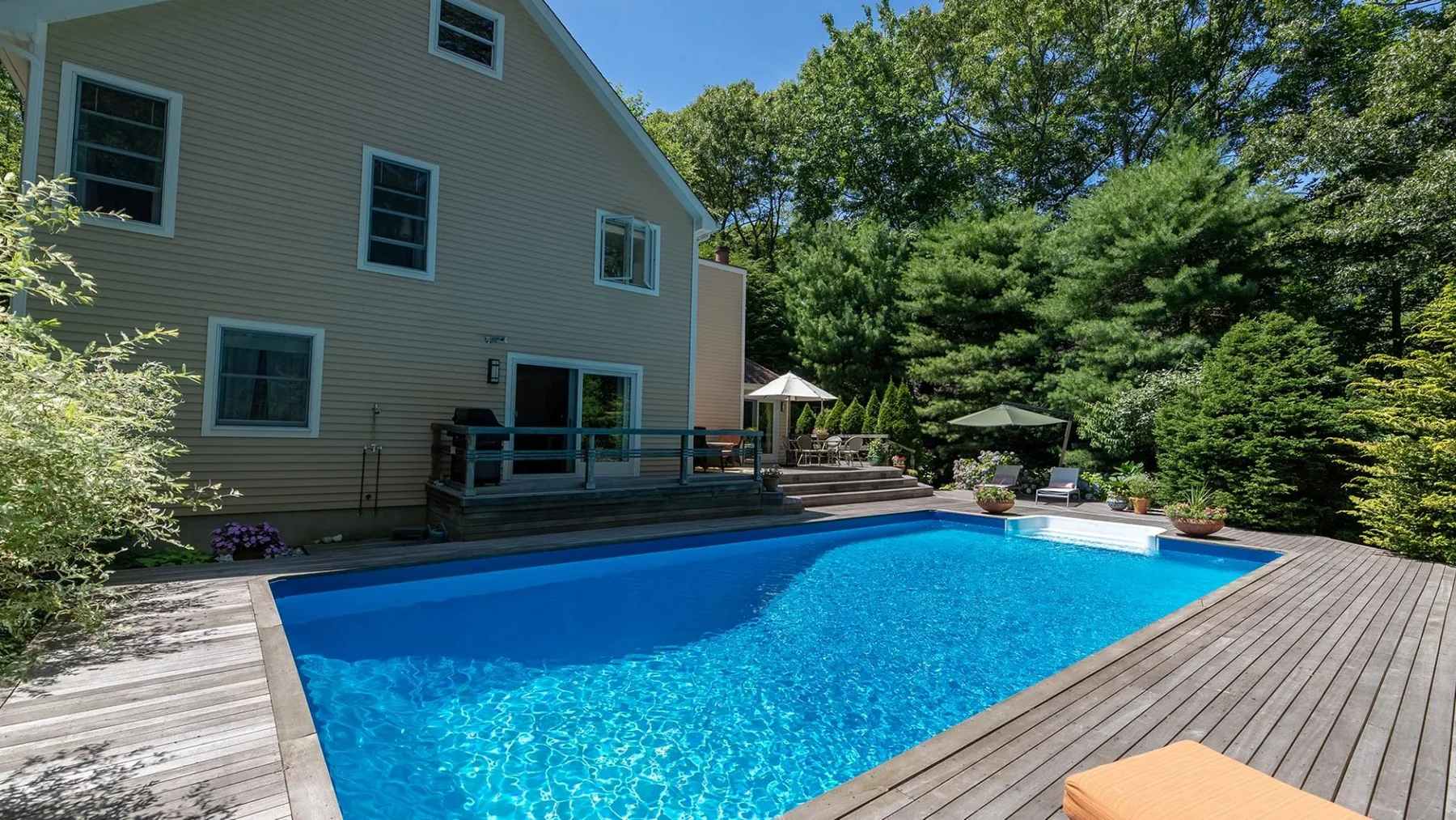 A swimming pool beside a house, with a wooden deck and surrounding trees.