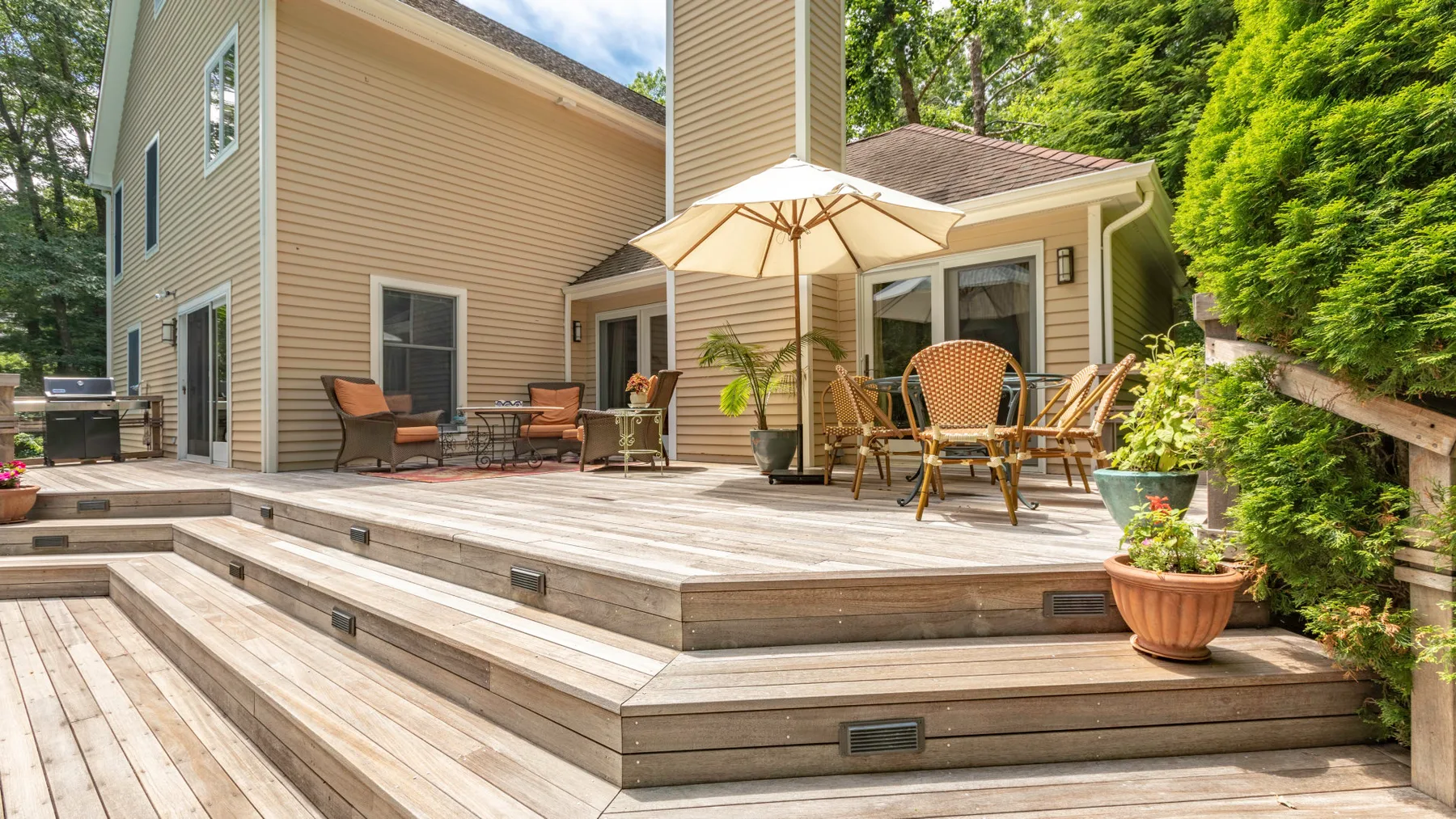 A raised wooden deck beside a beige house has outdoor seating and a patio umbrella.
