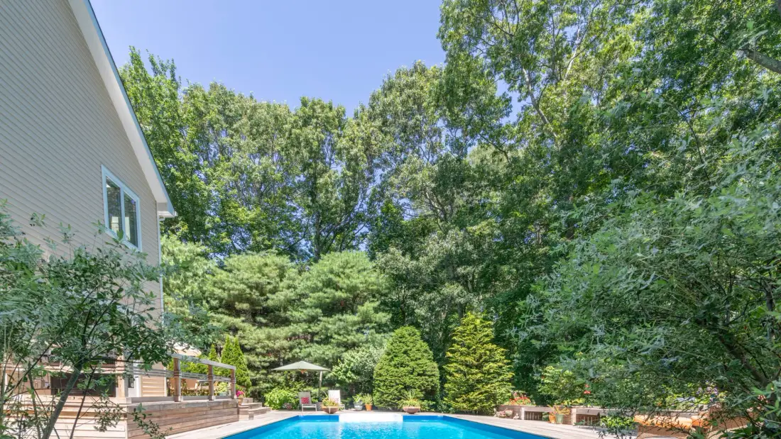 An outdoor swimming pool sits beside a house, surrounded by trees and greenery.