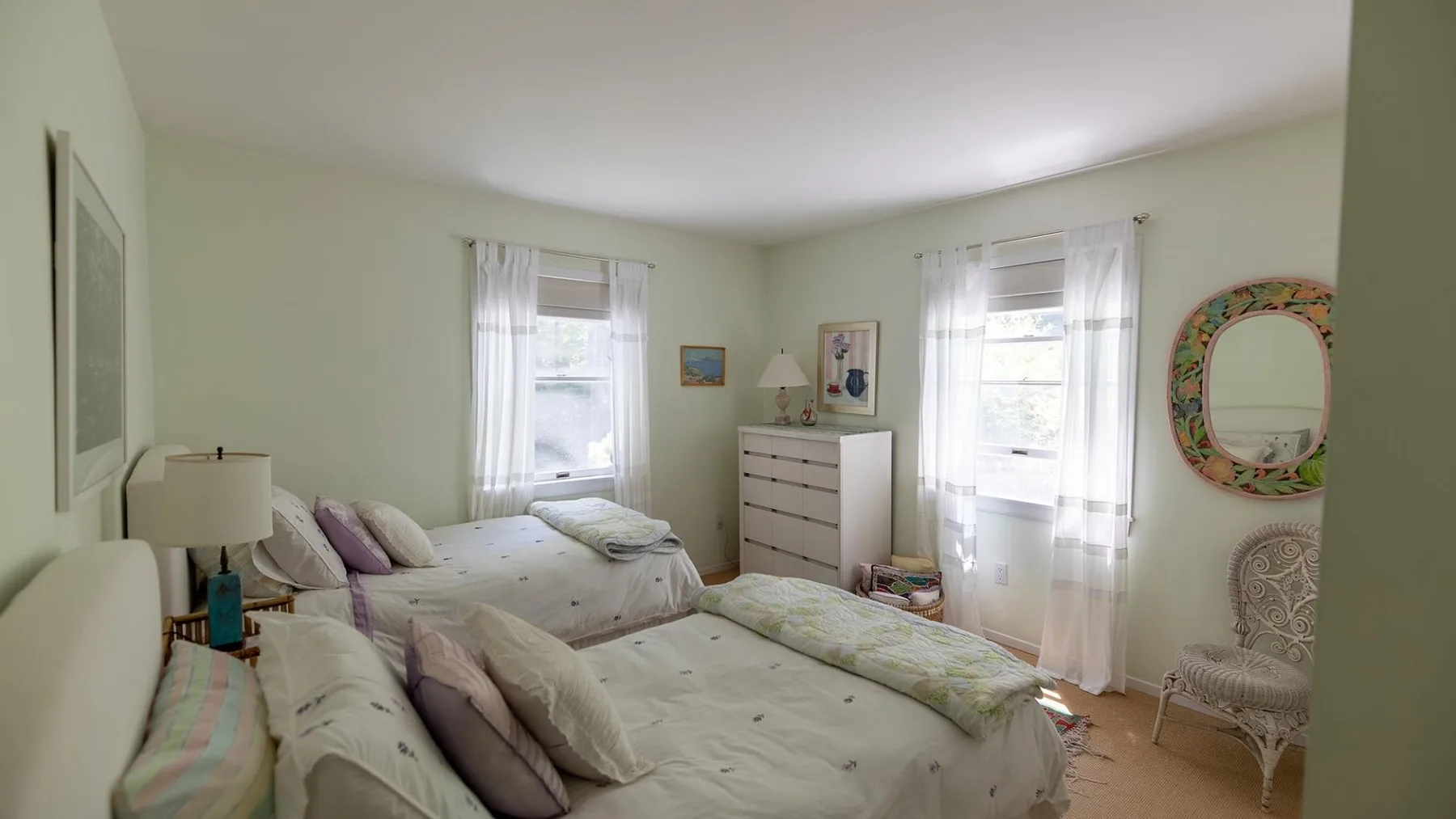 A bright bedroom with two beds, pale bedding, and windows with sheer curtains.