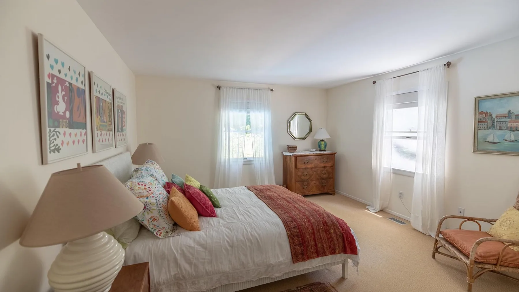 A bedroom with a bed, framed artwork, and windows with sheer curtains.