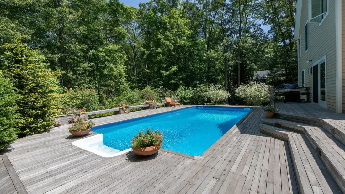 An outdoor swimming pool sits beside a wooden deck and leafy trees.