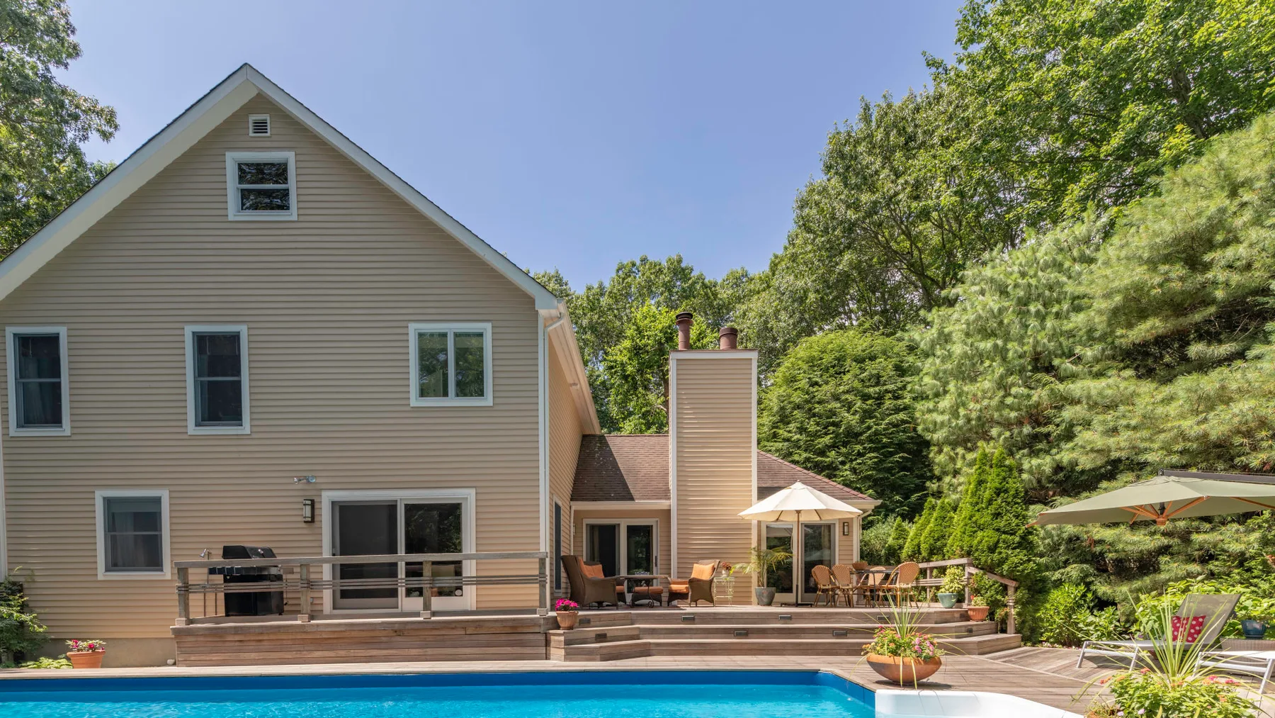 A beige house opens onto a deck and swimming pool, surrounded by trees.
