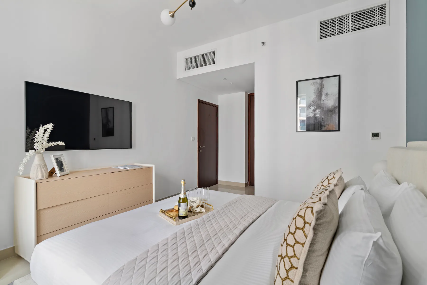Un dormitorio luminoso con una cama bien tendida, una cómoda y un televisor de pared.
