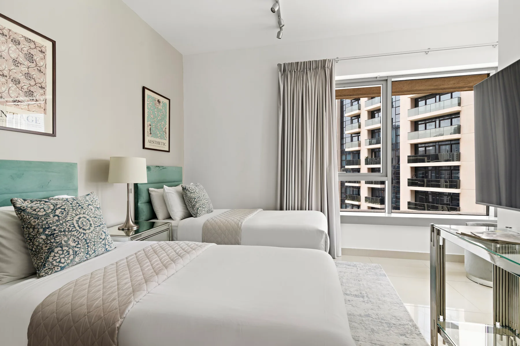 Un dormitorio luminoso con dos camas y grandes ventanas con vistas a edificios cercanos.