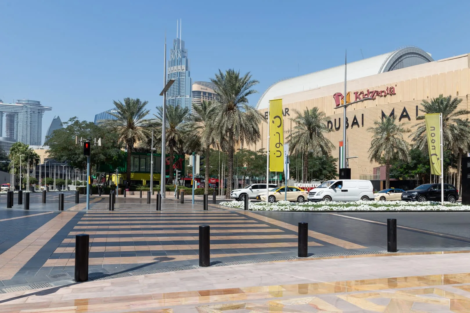 Una plaza peatonal pavimentada con palmeras y un gran centro comercial al fondo.