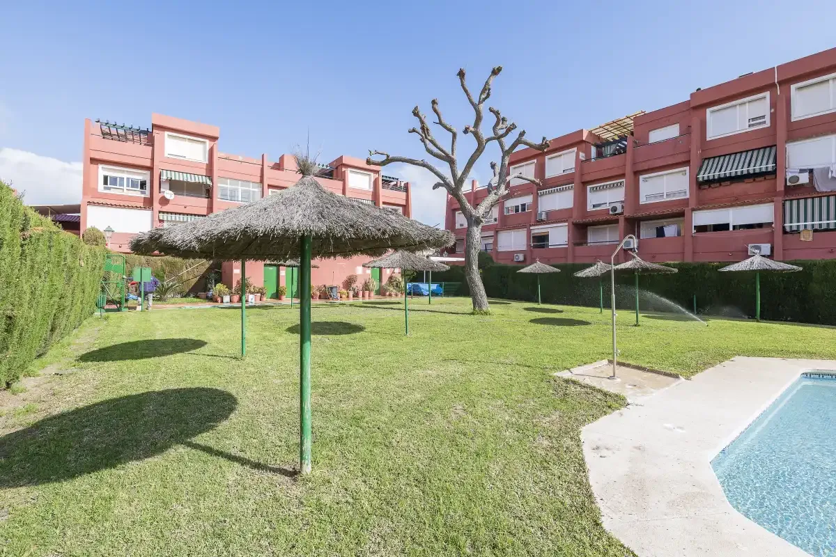 A grassy courtyard with shade umbrellas beside a swimming pool and apartment buildings.