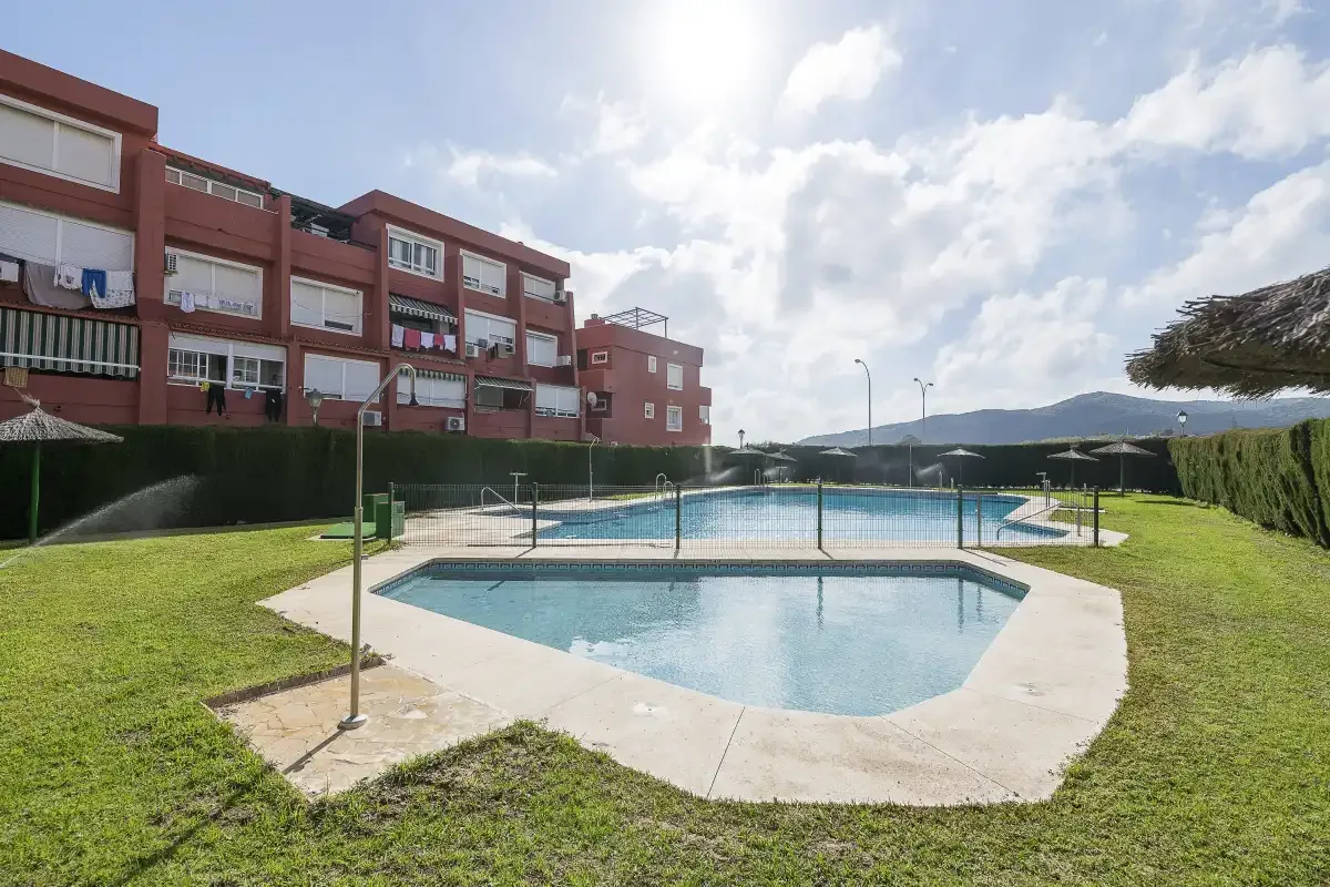 An outdoor swimming pool sits on a lawn beside a red apartment building.