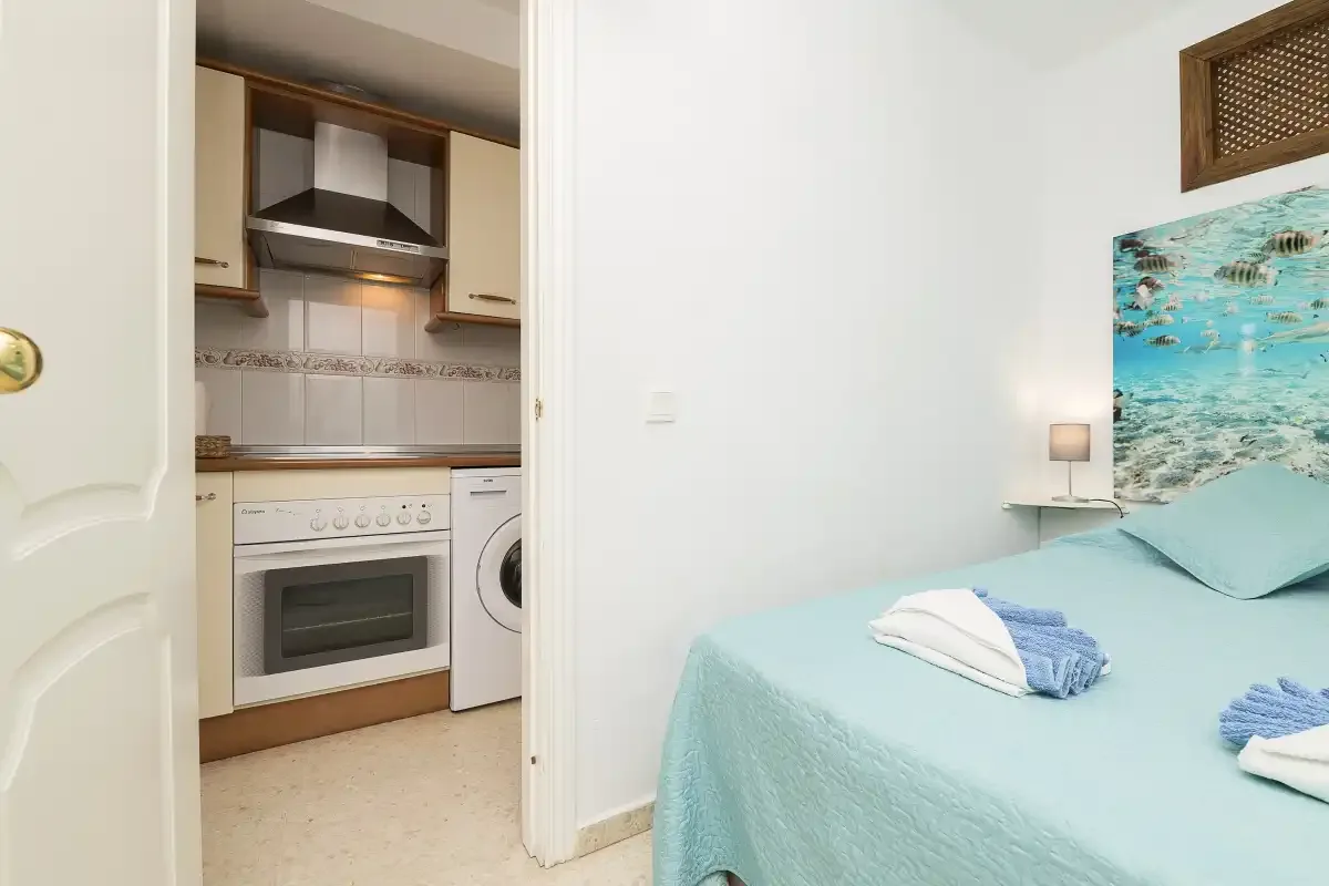 A bedroom with a turquoise bed beside a compact kitchen.