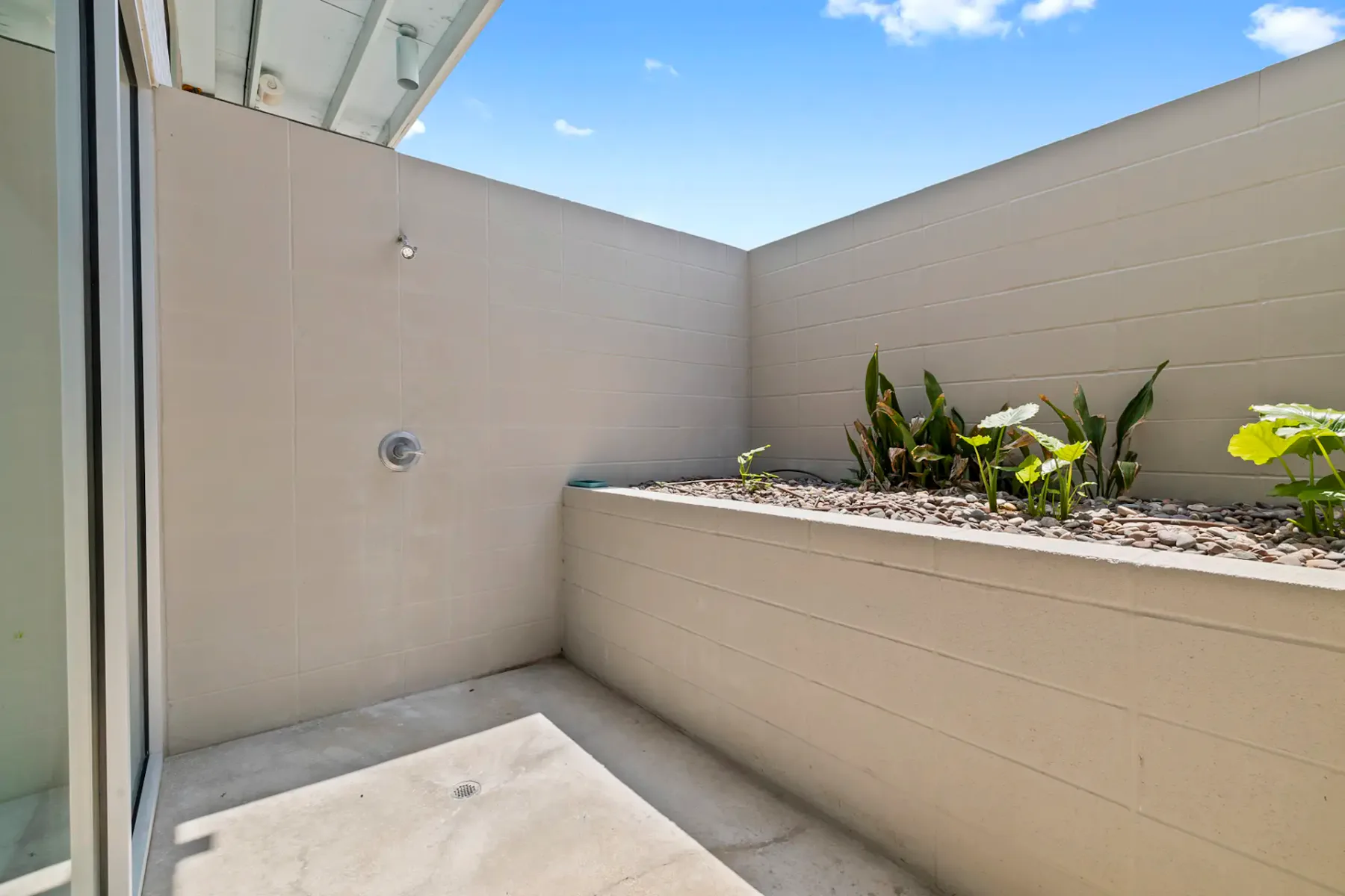 Zona de ducha exterior minimalista con suelo de hormigón, paredes beige y una pequeña jardinera con plantas verdes.