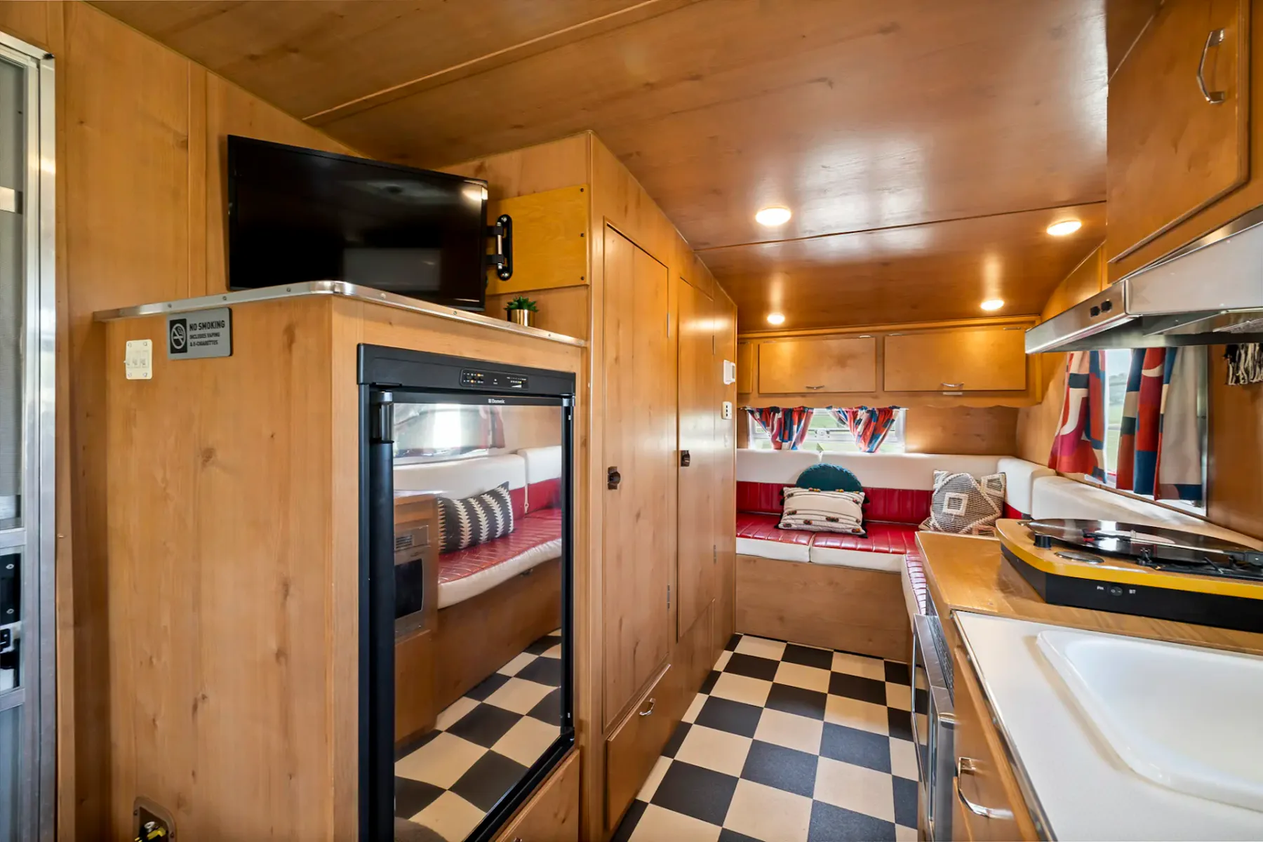 Cocina revestida de madera con encimeras blancas, fregadero, cafetera, tostadora, especiero y cortinas con estampado rojo. Pequeña placa de cocina sobre la encimera.