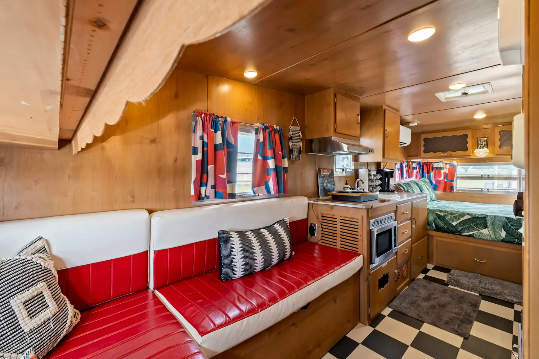 Interior de una caravana de época con asientos rojos y blancos, suelo de cuadros blancos y negros, una pequeña cocina y una acogedora zona para dormir.