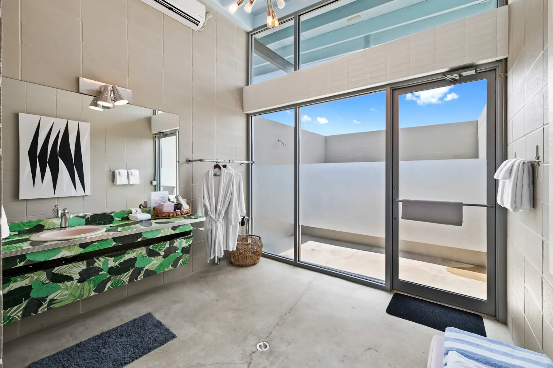 Baño moderno con lavabo de estampado tropical, albornoces en ganchos y una ventana grande. Decoración minimalista con arte abstracto y tonos neutros.