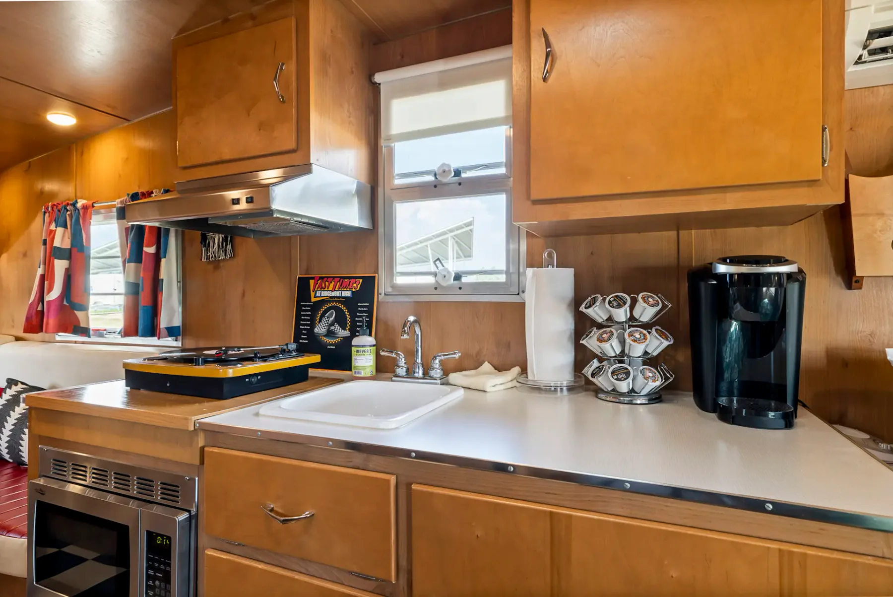Cocina revestida de madera con encimeras blancas, fregadero, cafetera, tostadora, especiero y cortinas con estampado rojo. Pequeña placa de cocina sobre la encimera.