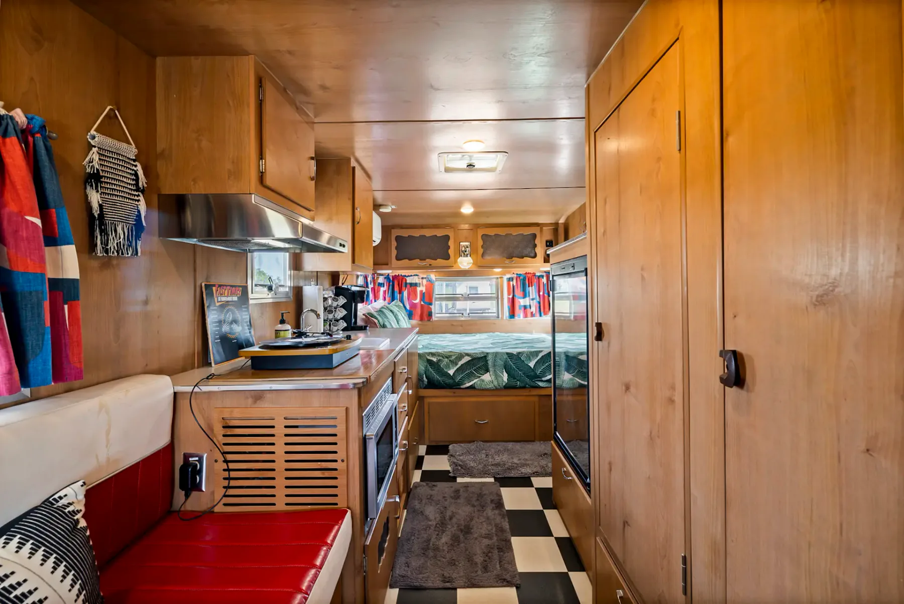 Interior de una caravana de época con asientos rojos y blancos, suelo de cuadros blancos y negros, una pequeña cocina y una acogedora zona para dormir.
