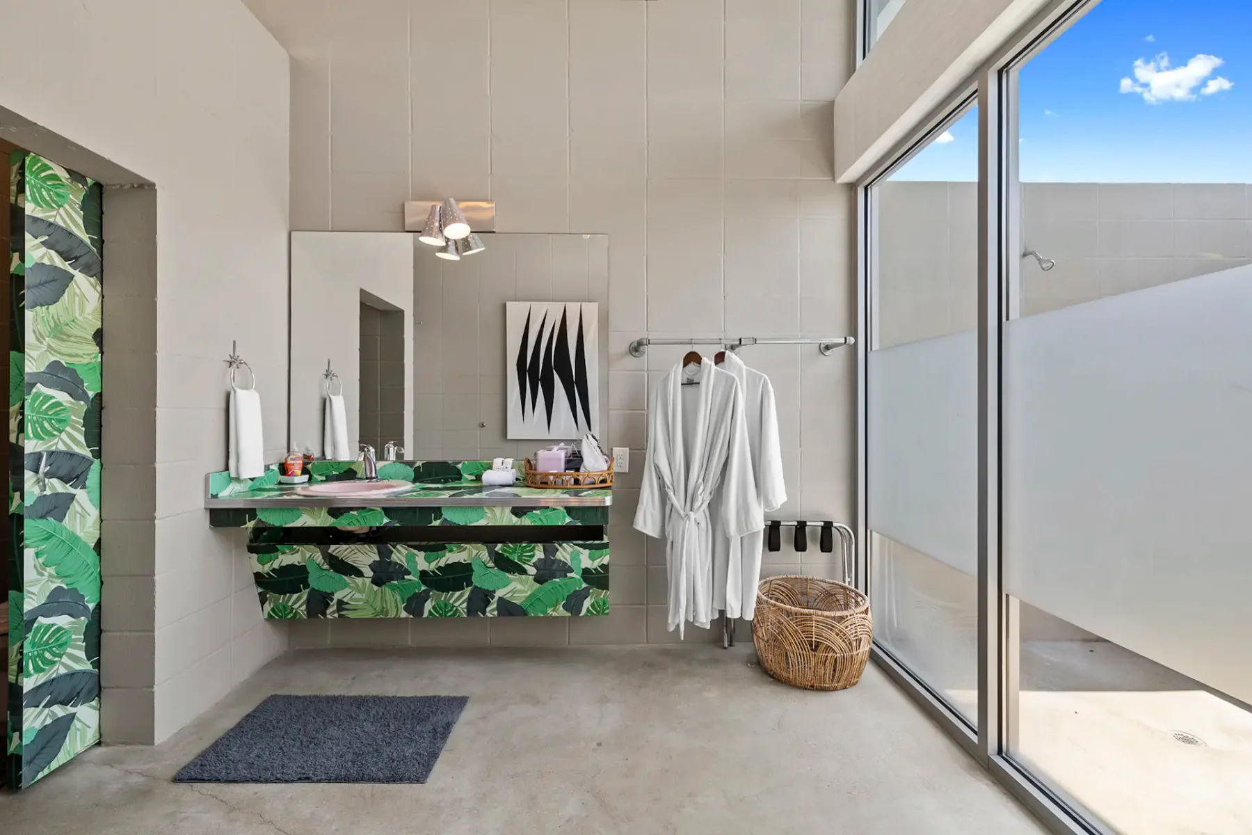 Baño moderno con lavabo de estampado tropical, albornoces en ganchos y una ventana grande. Decoración minimalista con arte abstracto y tonos neutros.