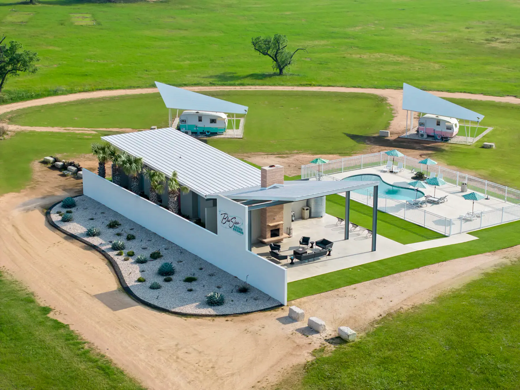 Casa moderna con piscina, rodeada de césped y caminos de tierra. Dos caravanas retro bajo toldos en las inmediaciones. Campo verde y frondoso al fondo.