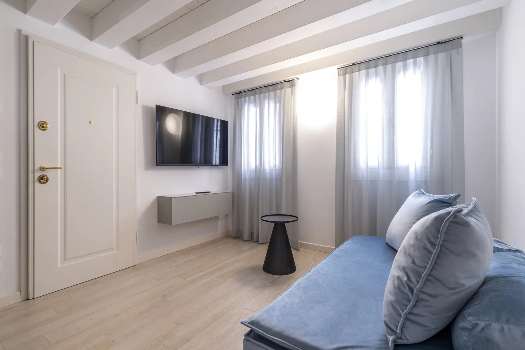 Una sala luminosa con asientos azules, cortinas translúcidas y un televisor en la pared.