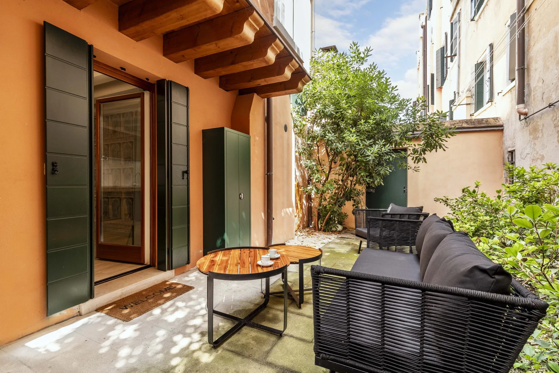 Un pequeño patio exterior con asientos, una mesa y plantas frondosas junto al edificio.