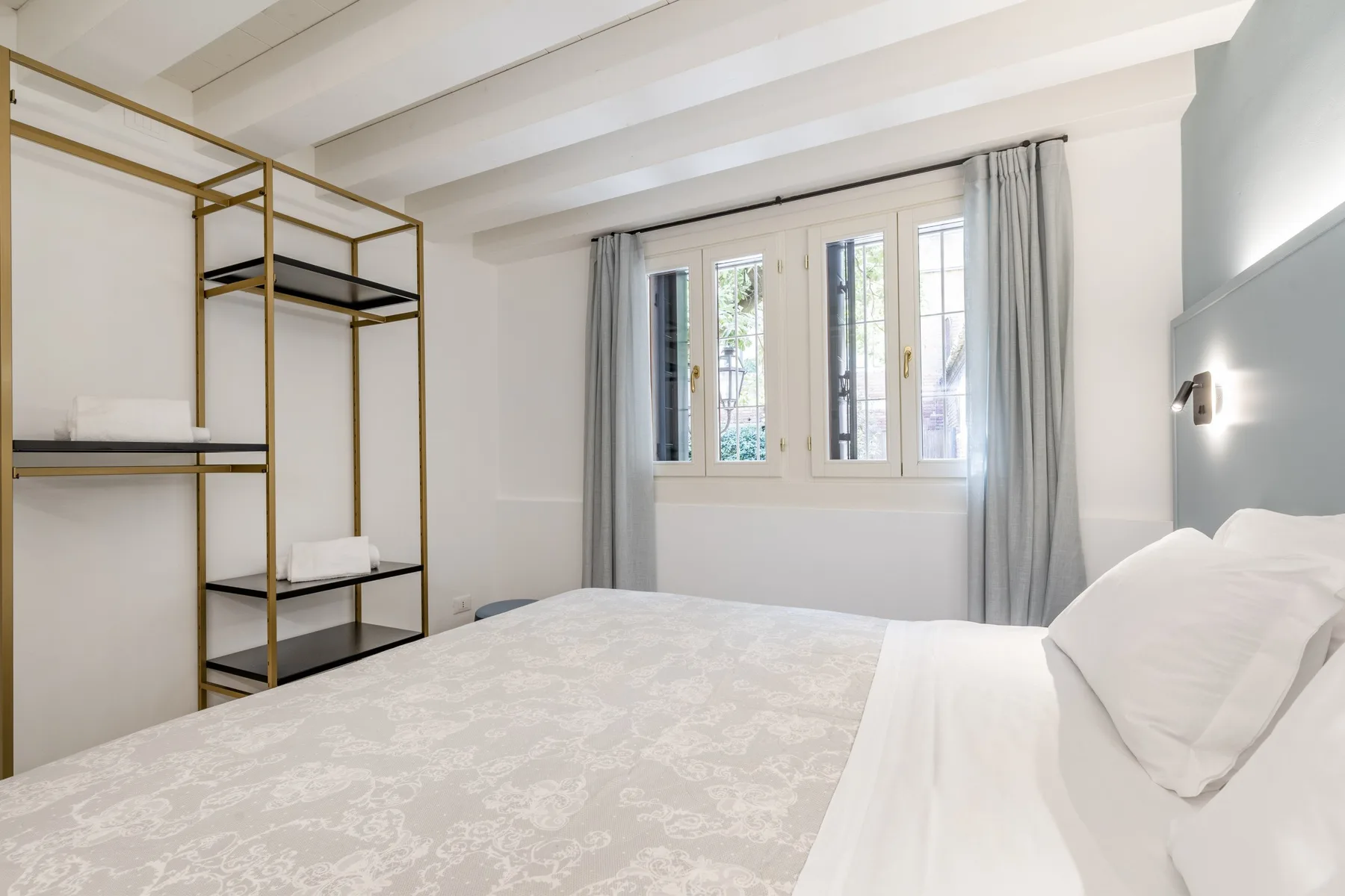 Un dormitorio luminoso con una cama, ventanas altas y un perchero abierto.