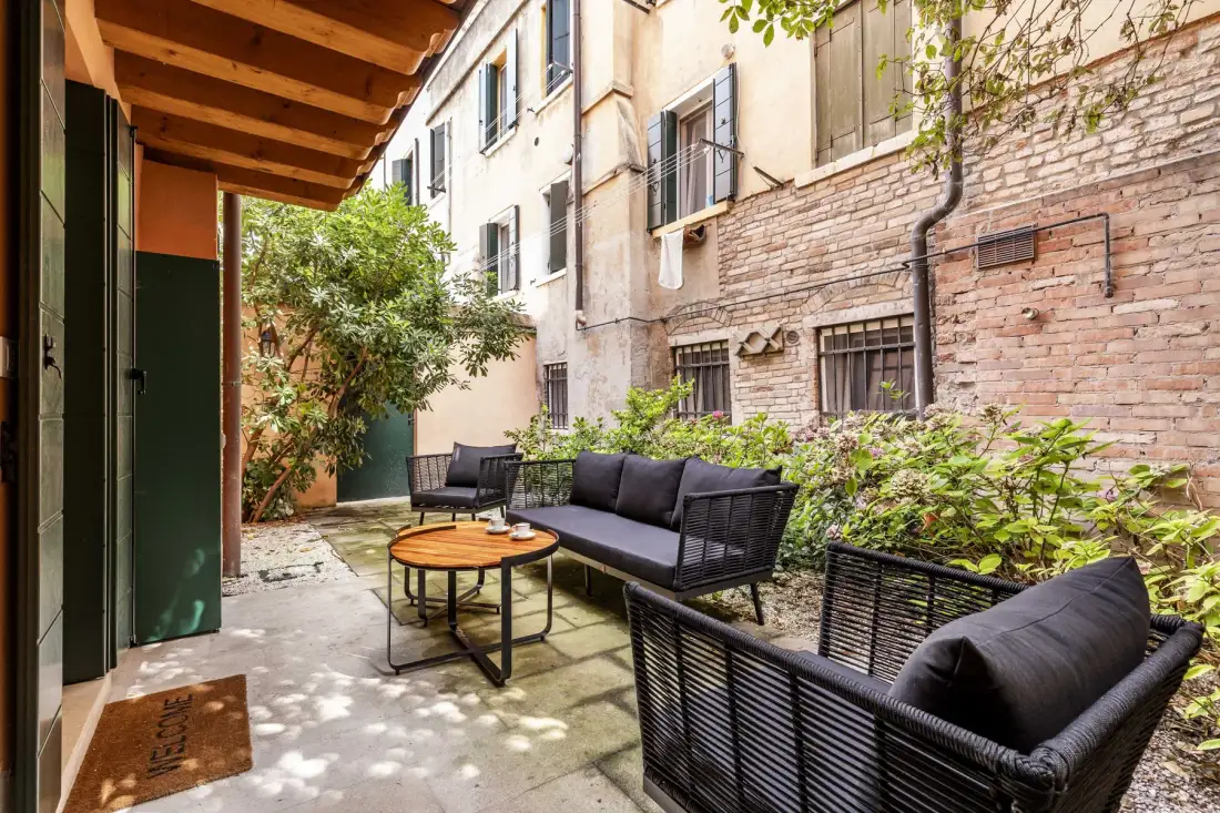 Zona de estar exterior pavimentada, con asientos oscuros y una mesa redonda junto a un muro de ladrillo.