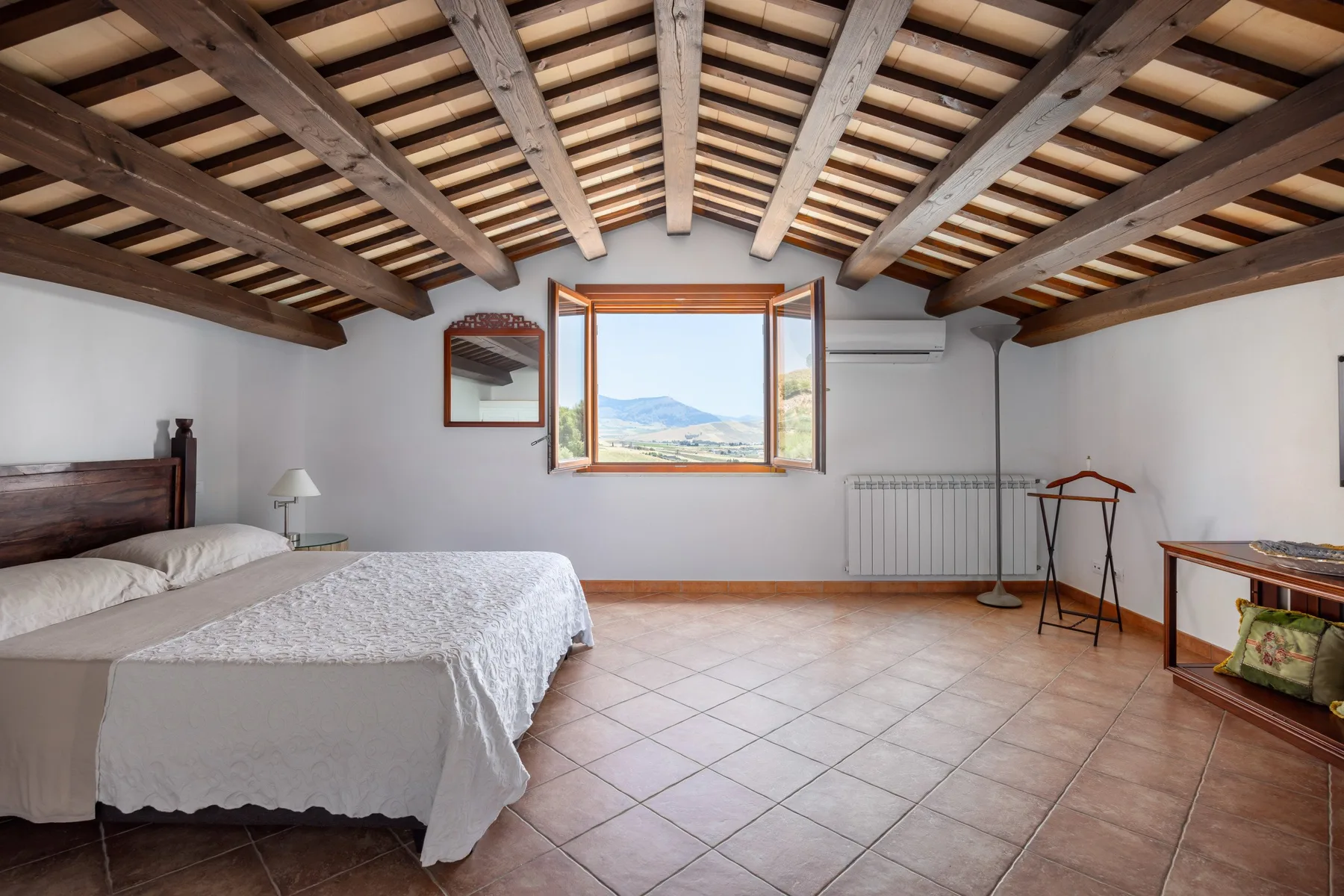 A bedroom with a bed beneath exposed ceiling beams and a window overlooking hills.