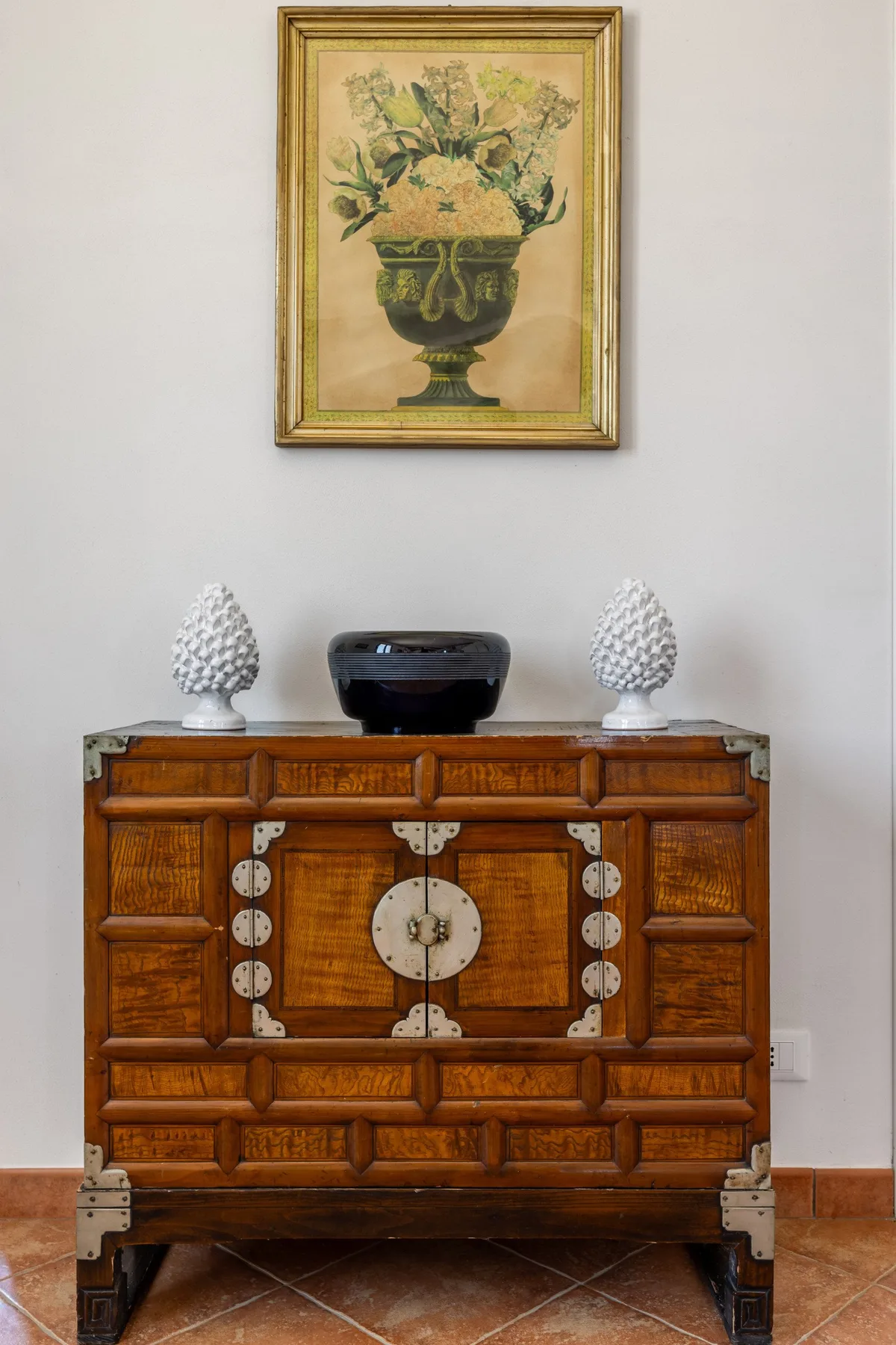 A wooden cabinet stands against a pale wall beneath a framed floral picture.