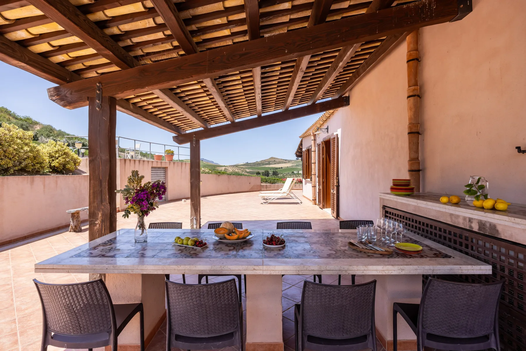 An outdoor dining area beneath a wooden pergola, with a long table and chairs.