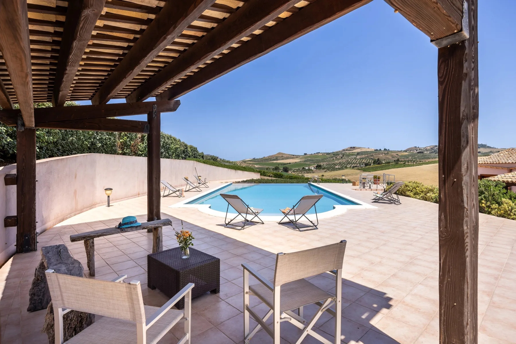 A tiled outdoor pool terrace with lounge seating beneath a wooden pergola.